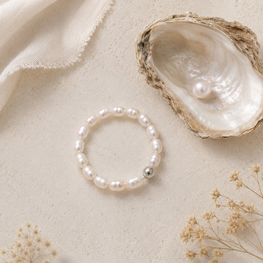 A beaded stretch bracelet made of white freshwater pearls with a single silver bead accent, displayed on a textured beige surface next to an oyster shell containing a pearl.