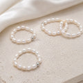 Four pearl beaded rings with small silver spacer beads, arranged on a textured beige stone surface with a draped white satin cloth in the background.