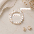 A pearl beaded bracelet with a single silver bead, displayed on a textured beige surface with decorative dried flowers and fabric. The image includes measurement callouts indicating the bracelet's diameter and circumference.