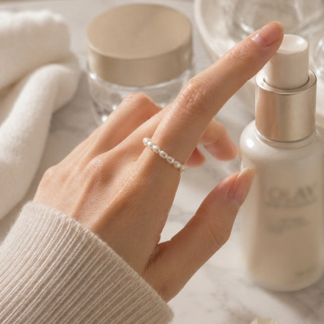 A close-up lifestyle shot of a hand wearing a delicate white pearl ring, reaching toward a skincare pump bottle on a marble surface with soft, warm lighting.